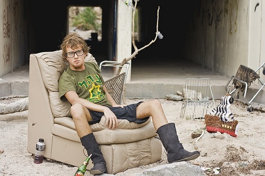 a picture of a young homeless man, sitting on a couch in an outdoor homeless camp